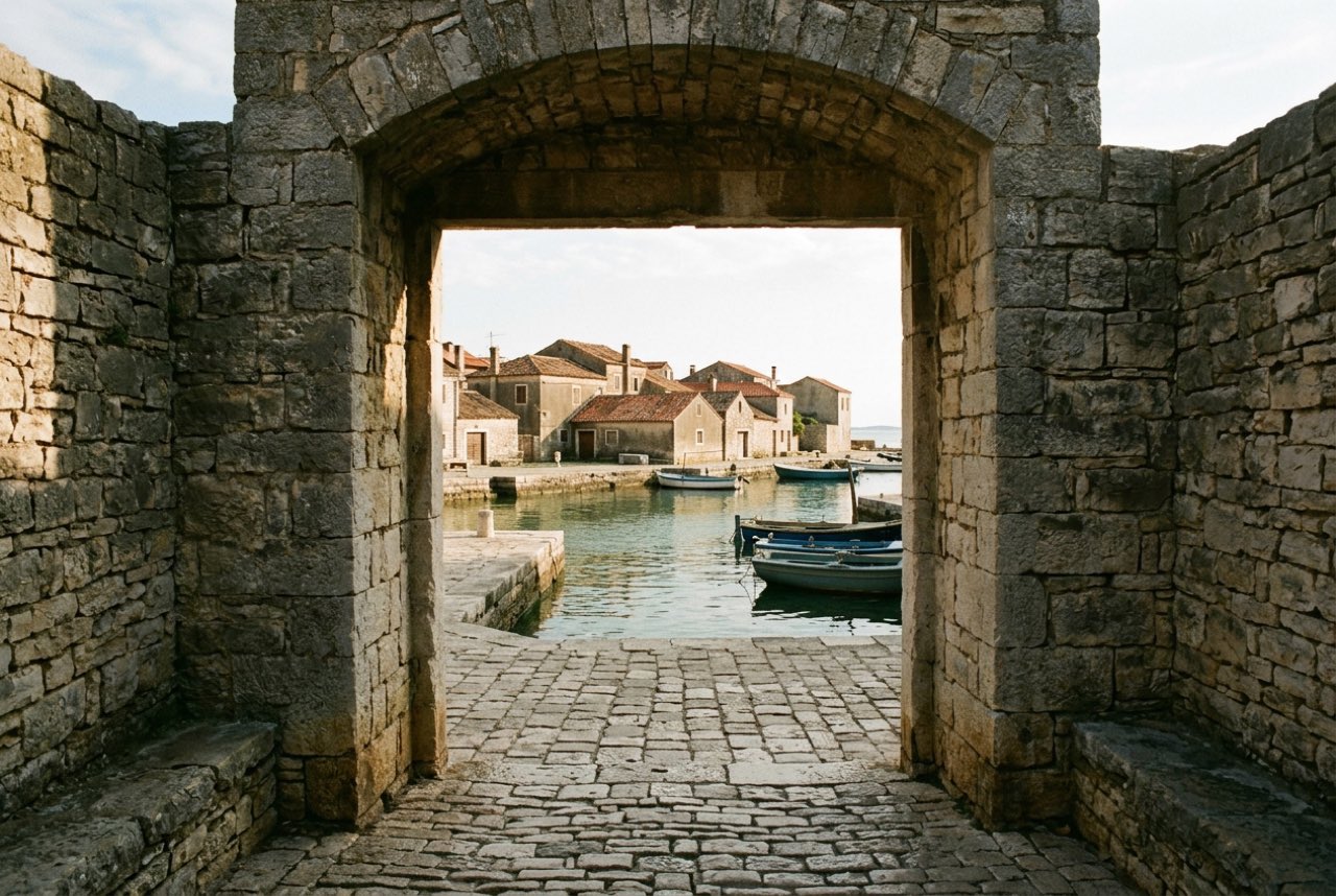A stone town gate opening onto a narrow harbour of clear green water and low tiled roofs, warm afternoon shadow