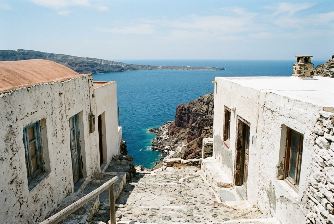 A gap between whitewashed cliff top houses opening onto a vertical drop to deep blue Aegean water, bare volcanic rock, hard clear light