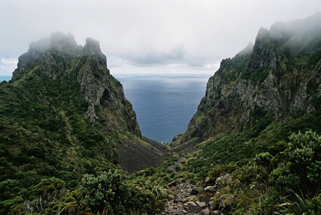 A cleft between two enormous green volcanic spires opening onto a deep Pacific bay, heavy cloud on the ridges, no vessels